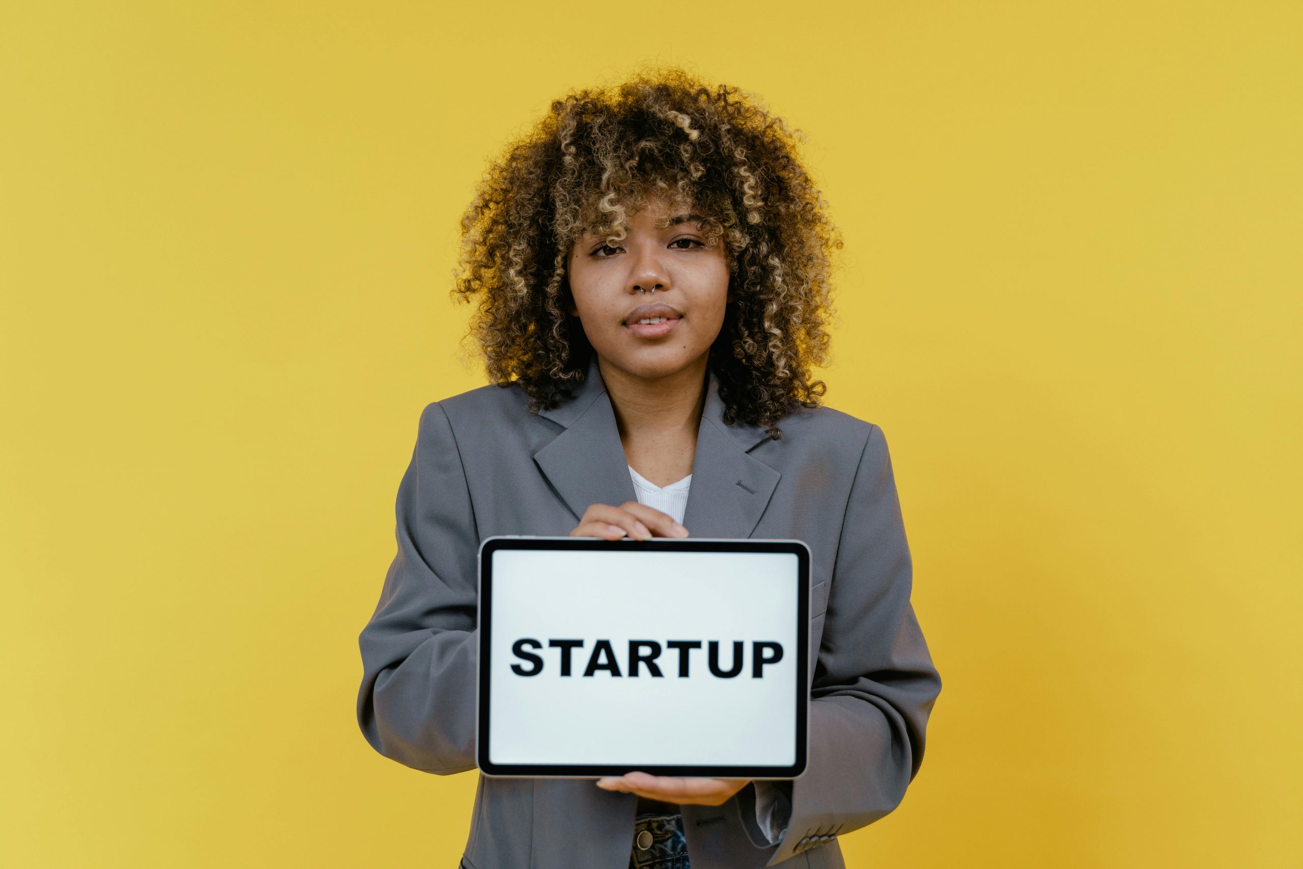 Portrait of a woman holding a tablet showing the word 'STARTUP' against a yellow background.